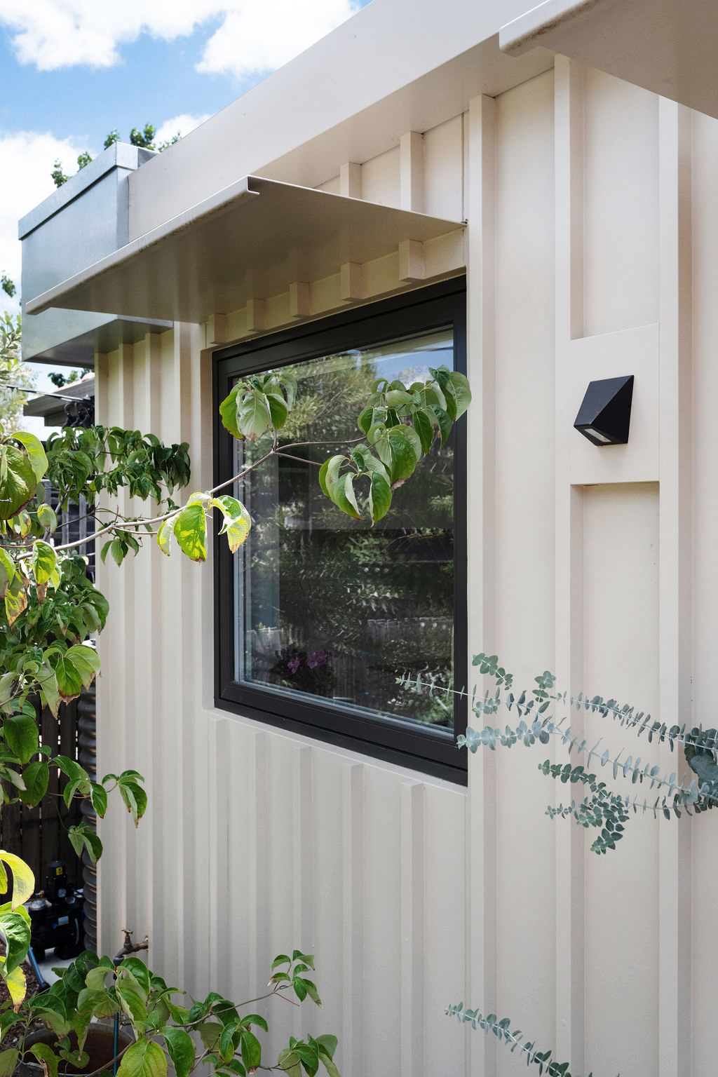 Cream‑coloured cladding with black‑framed window and metal awning surrounded by greenery